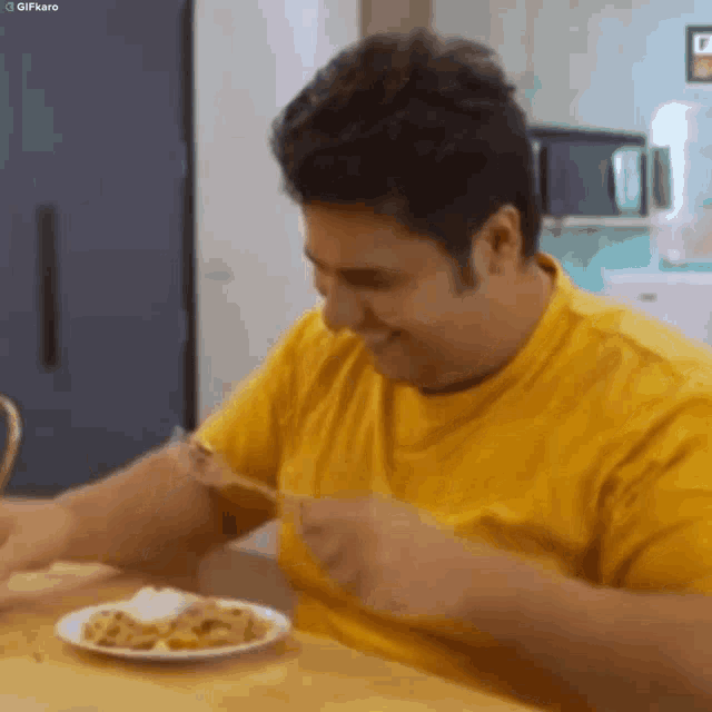 a man in a yellow shirt is sitting at a table eating a plate of food with a fork and knife .