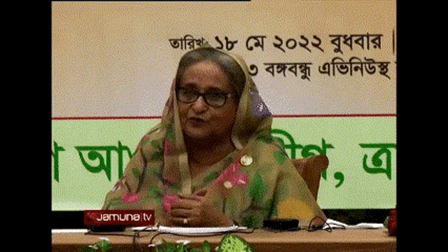 a woman wearing glasses sits at a table in front of a sign that says ' jamuna tv ' on it