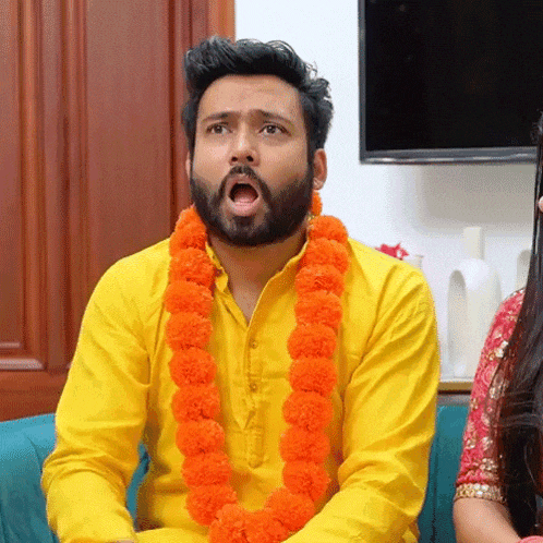 a man with a beard wearing a yellow shirt and an orange garland around his neck is sitting on a couch .