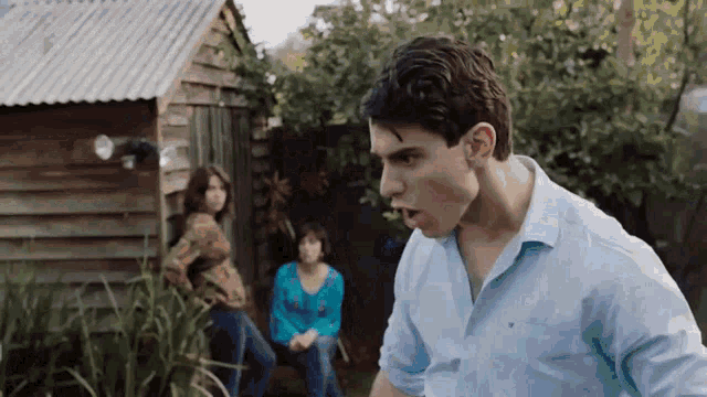 a man in a blue shirt is standing in front of a wooden shed while a woman sits on a bench .