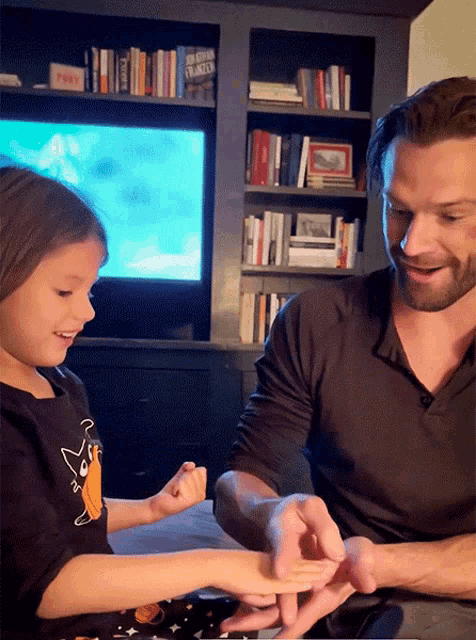 a man and a little girl are playing with their hands and the little girl is wearing a black shirt with a cat on the front