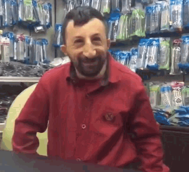 a man with a beard wearing a red shirt is standing in front of a shelf of remote controls .