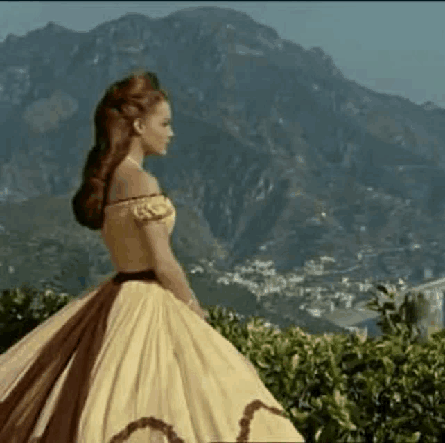 a woman in a yellow dress is walking in front of a mountain .