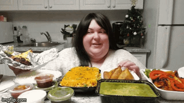 a woman is sitting at a table with a lot of food and a christmas tree in the background ..