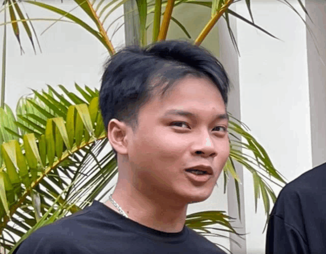 a young man wearing a black shirt and a chain around his neck stands in front of a palm tree