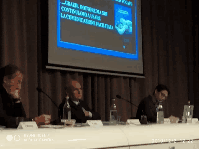 three men sit at a table with microphones in front of a screen that says grazie