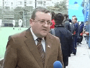 a man in a suit and tie is standing in front of a soccer field talking into a microphone ..