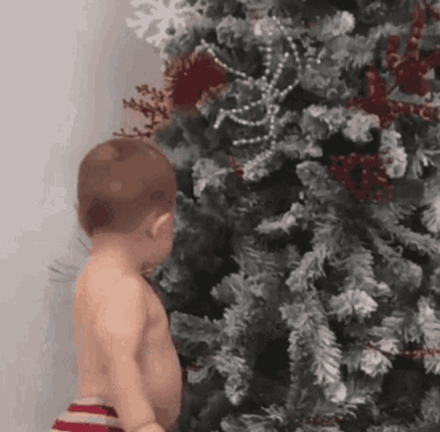 a shirtless baby standing in front of a christmas tree