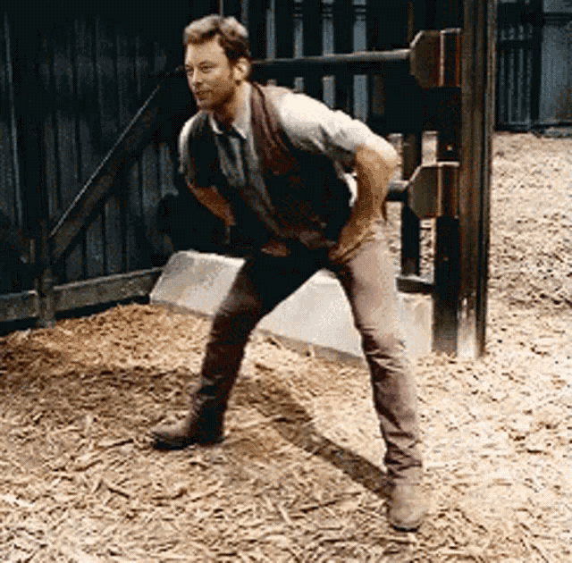 a man in a vest and brown pants is standing on one leg in front of a wooden fence .