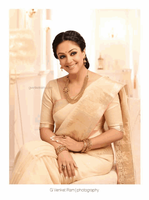 a woman wearing a white saree and gold jewelry is sitting on a couch