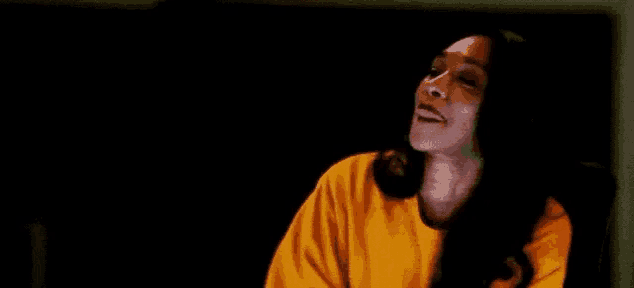 a woman in a yellow shirt is sitting in a dark room and making a funny face .