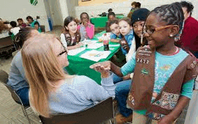 a girl scout is talking to a woman at a table while a group of girls are sitting at tables .