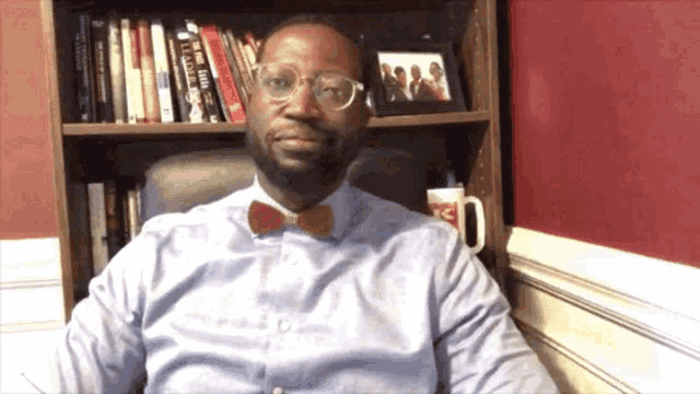 a man wearing glasses and a bow tie is sitting in front of a bookshelf with a book called leadership