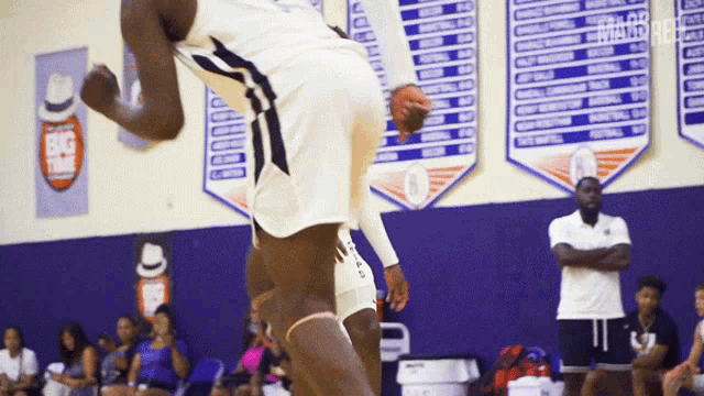 a basketball player is jumping in the air in front of a purple wall with a banner that says ' basketball ' on it