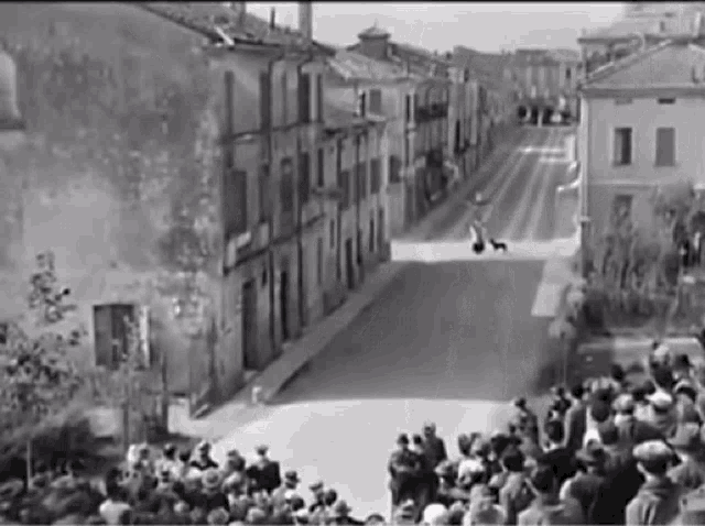 a black and white photo of a crowd of people watching a man walking down a street with a dog