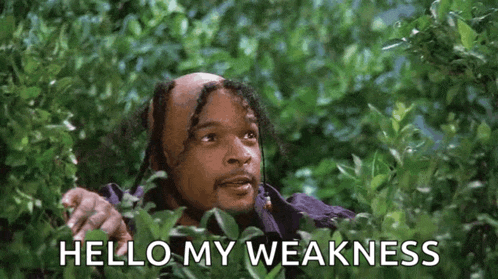 a bald man with dreadlocks is peeking out from behind a bush and saying `` hello my weakness '' .