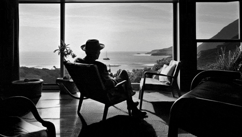 a man in a hat sits in a rocking chair looking out a window at the ocean