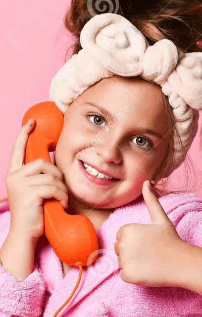 a little girl wearing a headband talking on an orange telephone and giving a thumbs up