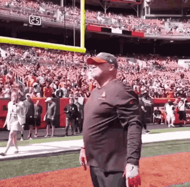 a man in a brown shirt is standing on a football field in front of a crowd with the number 25 on the scoreboard .