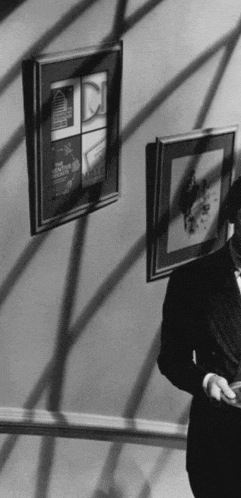 a black and white photo of a man standing in front of a wall with framed pictures on it including one that says milles