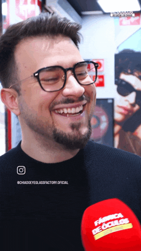 a man wearing glasses is smiling in front of a microphone that says fabrica de oculos on it