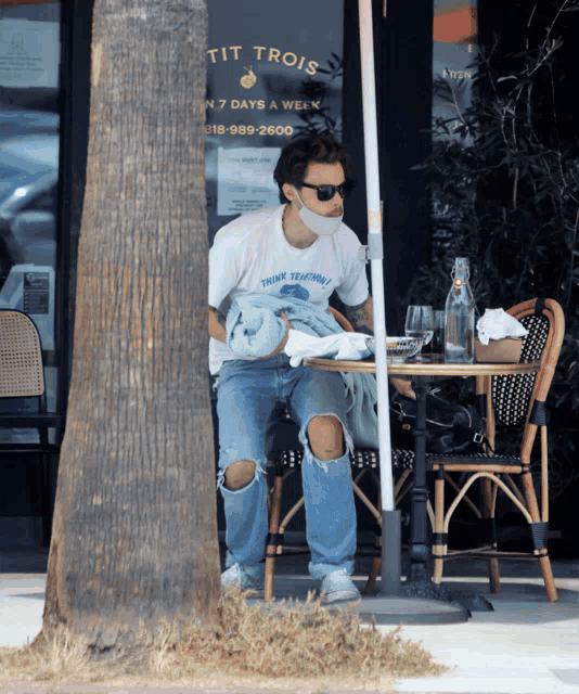 a man wearing a mask sits at a table in front of a restaurant called tit trois