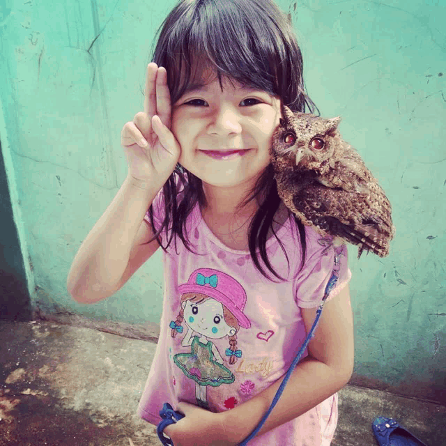 a little girl wearing a pink shirt with a girl on it is holding an owl on her shoulder