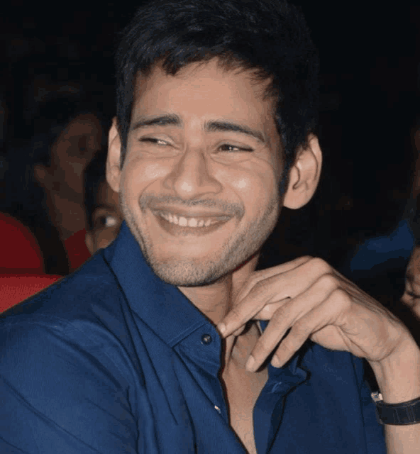 a man in a blue shirt smiles with his hand on his neck