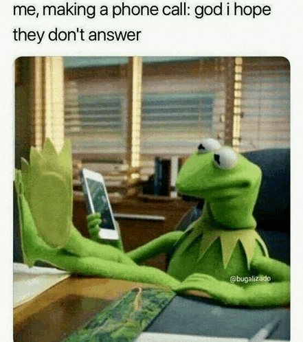 kermit the frog is sitting at a desk holding a cell phone and making a phone call .