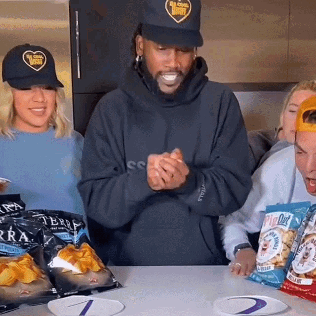 a man wearing a black hat with a heart on it stands in front of a table full of bags of popcorn