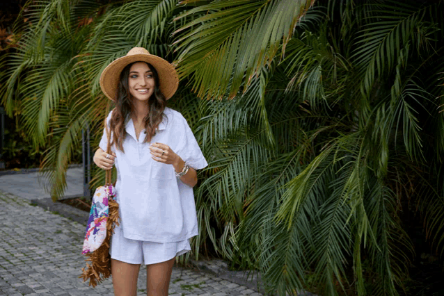 a woman wearing a straw hat and a white shirt is standing in front of palm trees