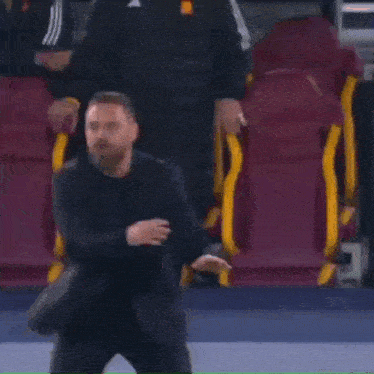 a man in a suit is standing in front of a row of red and yellow seats on a soccer field .