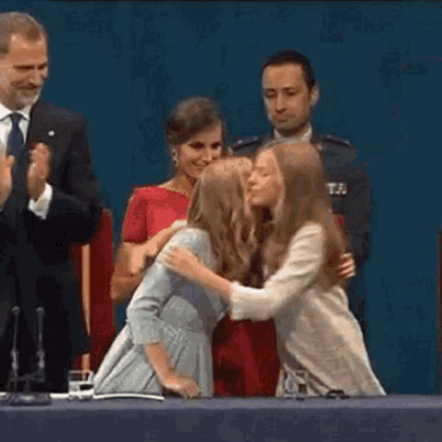 two young girls are hugging each other while sitting at a table in front of a man in a suit and tie .