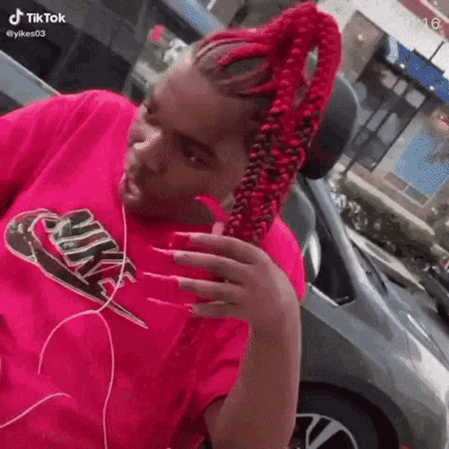 a woman with red braids and long nails wearing a pink nike shirt is standing in front of a car .