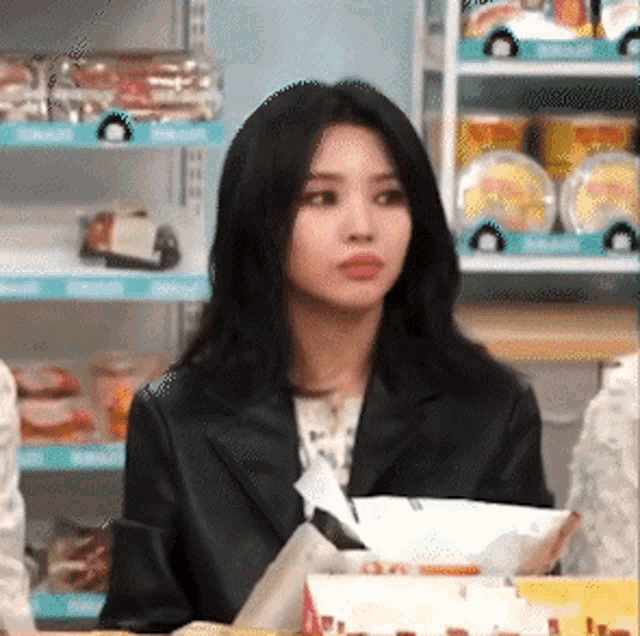 a woman in a black jacket is sitting at a table in front of a shelf with food on it .