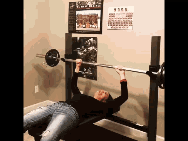 a woman is lifting a barbell on a bench in front of a poster that says " we make marines "