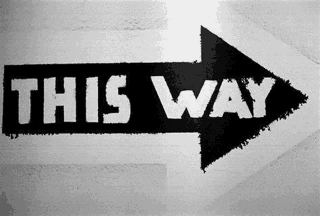 a black and white photo of an arrow pointing to the right with the words `` this way '' painted on it .