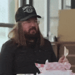 a man with long hair and a beard is sitting at a table eating food with a fork and knife .