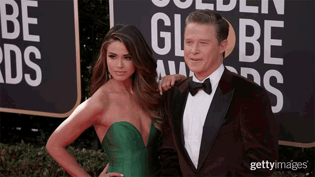 a man in a tuxedo and a woman in a green dress pose on a red carpet in front of a golden globe sign