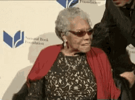 an elderly woman wearing sunglasses and a red scarf is sitting in front of a wall that says national book foundation