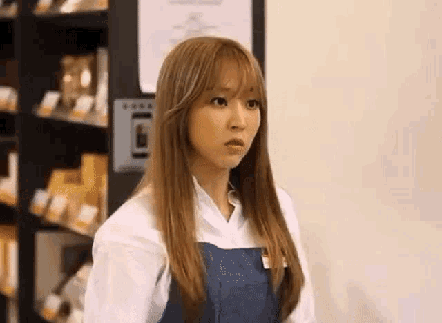 a woman in a white shirt and blue apron is standing in front of a shelf in a store .