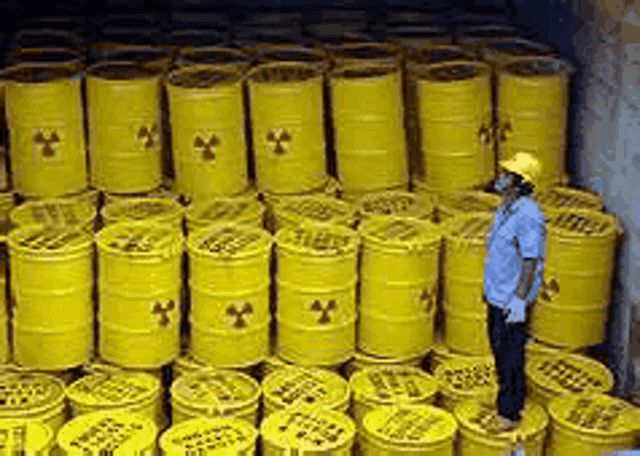 a man in a hard hat is standing in front of a pile of yellow barrels with radioactive symbols on them .