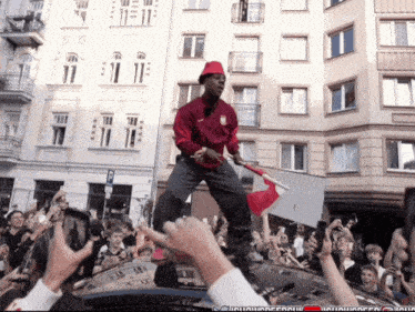 a man in a red jacket stands on top of a car holding a flag in front of a crowd