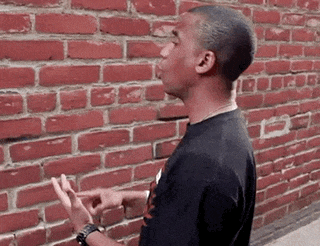 a man in a black shirt is standing in front of a red brick wall making a funny face .