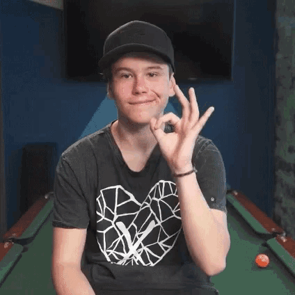 a young man wearing a hat and a black shirt is sitting in front of a pool table giving an ok sign .