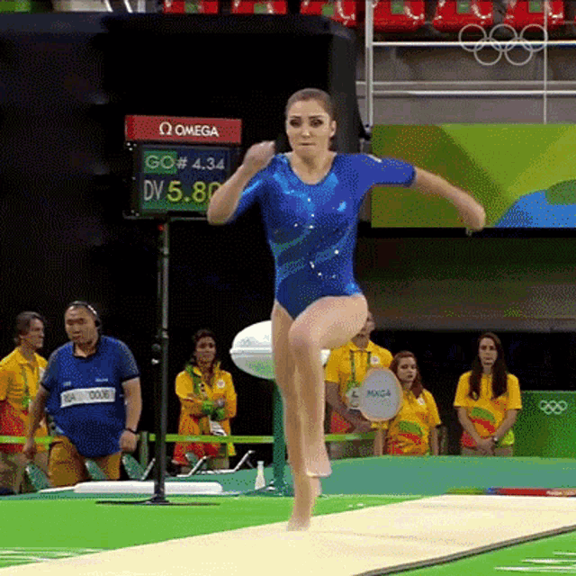 a woman in a blue leotard is doing a trick on a trampoline with a omega sign in the background