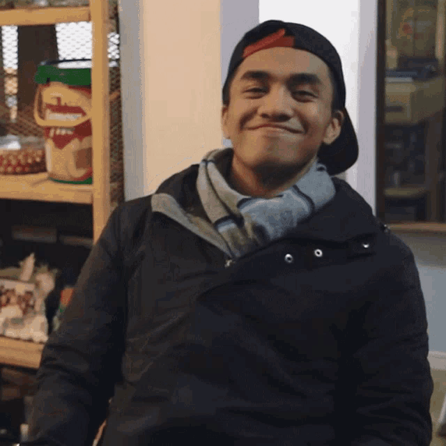 a man wearing a hat and a scarf smiles in front of a shelf with a bucket of beans on it