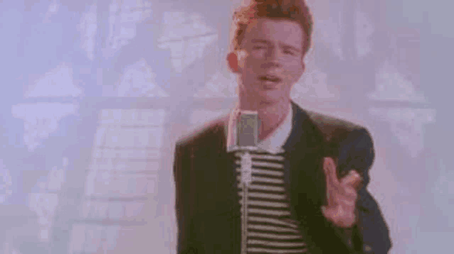 rick astley is singing into a microphone in front of a wall .