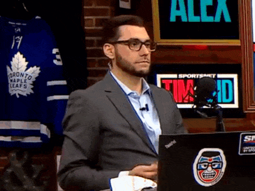 a man in a suit is sitting in front of a laptop with a toronto maple leafs jersey on the wall behind him