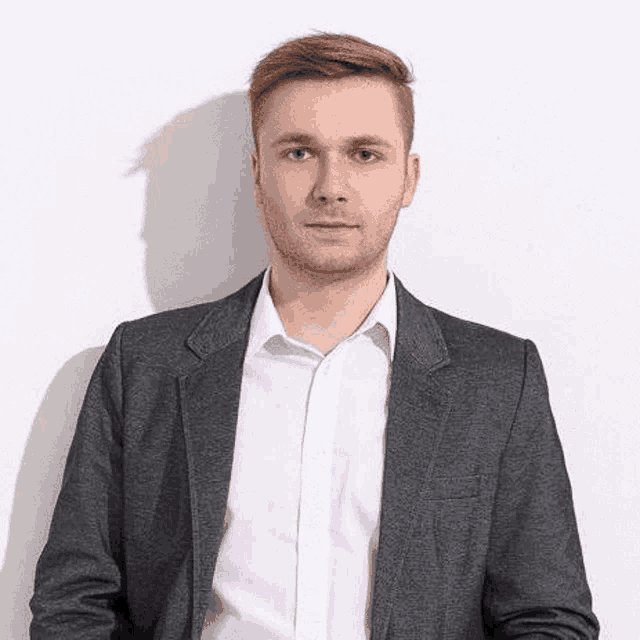 a man in a suit and white shirt is standing in front of a white wall and looking at the camera .